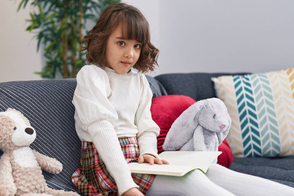 Adorable hispanic girl reading book sitting on sofa at home - Foto, afbeelding