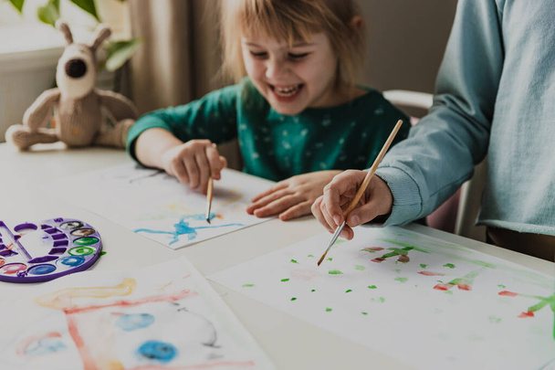 Two cute little sisters or classmates are painting on table with brush. Small girls are laughing. Kids are in kindergarten or at home. Child in living room. Home schooling concept, doing homework  - 写真・画像