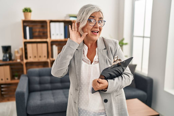 Frau mittleren Alters mit grauen Haaren im Beratungsbüro lächelt mit der Hand über dem Ohr und lauscht Gerüchten oder Klatsch. Konzept für Gehörlosigkeit.  - Foto, Bild