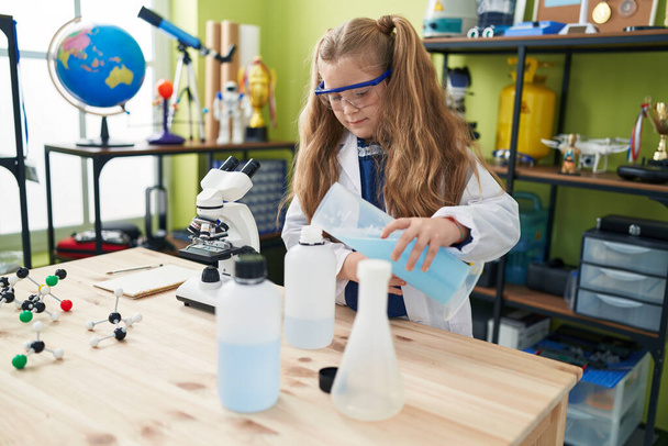 Adorable blonde girl student pouring liquid on bottle at laboratory classroom - Foto, imagen