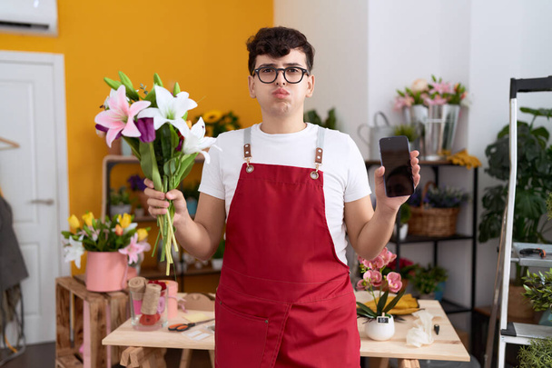 Young non binary man working at florist shop showing smartphone screen puffing cheeks with funny face. mouth inflated with air, catching air.  - Valokuva, kuva