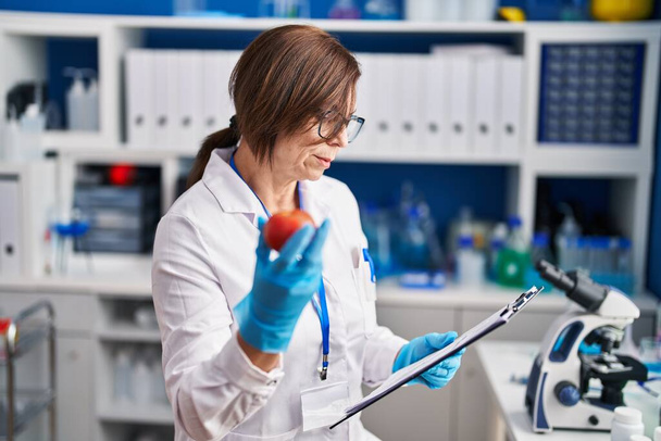 Middelbare leeftijd vrouw wetenschapper met appel lezen rapport in laboratorium - Foto, afbeelding