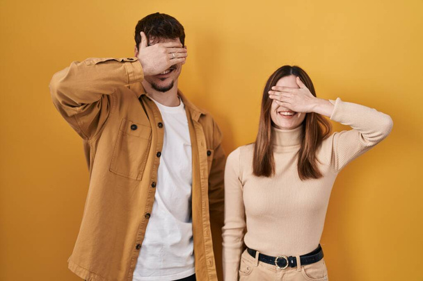 Young hispanic couple standing over yellow background smiling and laughing with hand on face covering eyes for surprise. blind concept.  - Fotó, kép