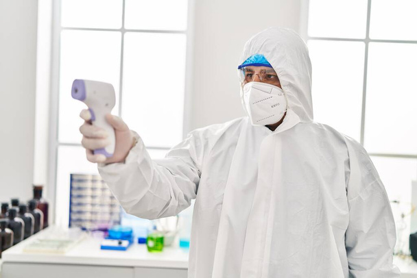Young latin man scientist wearing covid protection uniform holding thermometer at laboratory - Photo, Image