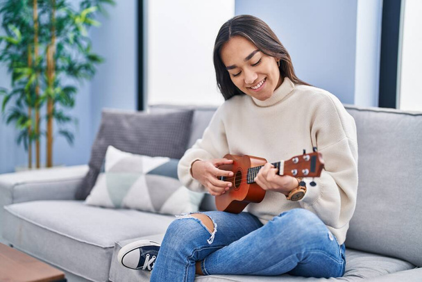 Молодая латиноамериканка, играющая на укулеле, сидит дома на диване - Фото, изображение