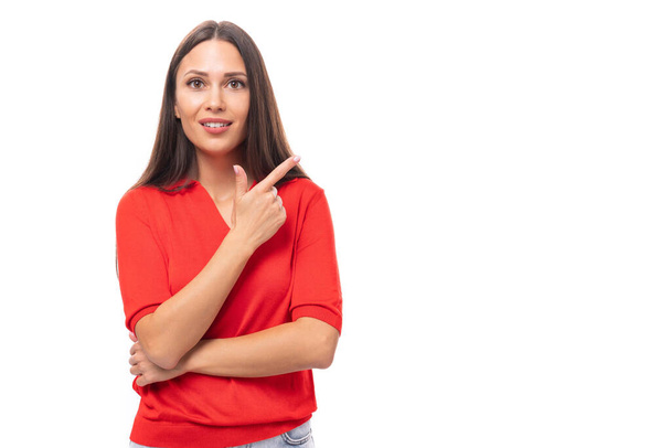 молодая симпатичная брюнетка кавказского цвета в красной рубашке с короткими рукавами указывает указательными пальцами на место для рекламы. - Фото, изображение