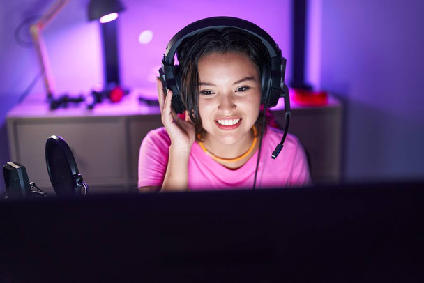 Молода іспаномовна жінка стример посміхається впевнено сидячи на столі в ігровій кімнаті
 - Фото, зображення