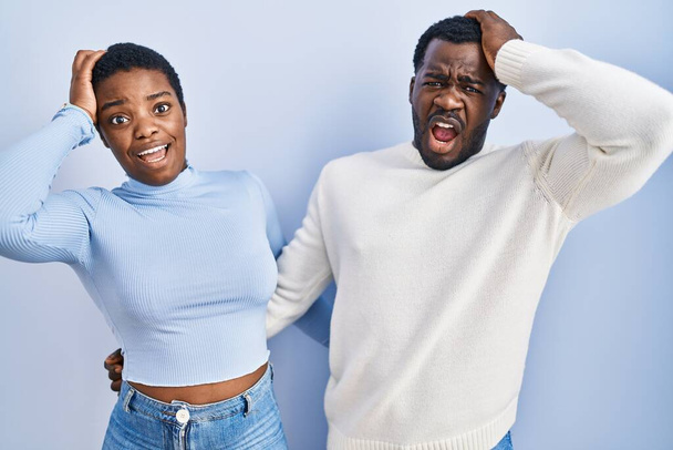 Junge afrikanisch-amerikanische Paar steht vor blauem Hintergrund verrückt und verängstigt mit Händen auf dem Kopf, Angst und überrascht von Schock mit offenem Mund  - Foto, Bild