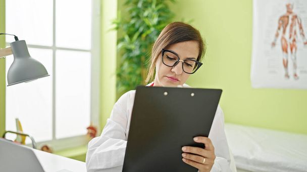 Middle eastern woman doctor reading clipboard thinking at clinic - Photo, Image