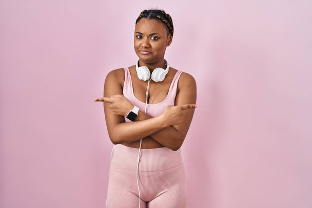Africká americká žena s copánky ve sportovním oblečení a sluchátky ukazujícími na obě strany prsty, odlišný směr nesouhlasí  - Fotografie, Obrázek