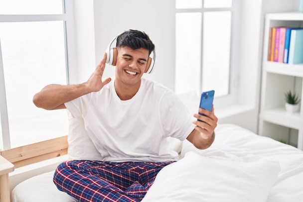 Joven hispano escuchando música sentado en la cama en el dormitorio - Foto, Imagen
