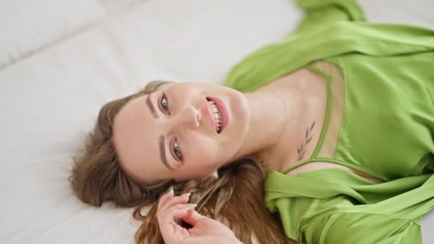 Νεαρή ξανθιά γυναίκα χαμογελά με αυτοπεποίθηση ξαπλωμένη στο κρεβάτι στο υπνοδωμάτιο - Πλάνα, βίντεο