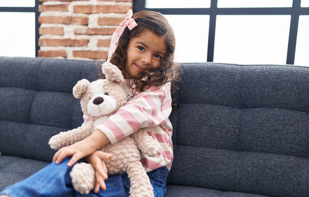 Αξιολάτρευτο ισπανόφωνο κορίτσι αγκαλιάζει αρκουδάκι κάθεται στον καναπέ στο σπίτι - Φωτογραφία, εικόνα