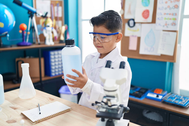 Adorable hispanic boy student smiling confident holding bottle with liquid at laboratory classroom - Foto, imagen