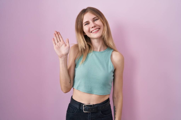 Блондинка-кавказка, стоящая на розовом фоне, отказывается от приветствия со словами "Привет, счастливый и улыбающийся, приветственный жест".  - Фото, изображение