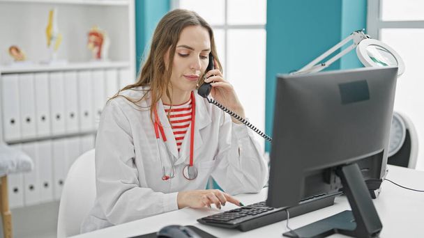 Nuori blondi nainen lääkäri käyttää tietokonetta puhuu puhelimessa klinikalla - Valokuva, kuva