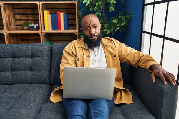 Afričan Američan pomocí notebooku doma sedí na pohovce myšlení postoj a střízlivý výraz vypadá sebevědomě  - Fotografie, Obrázek