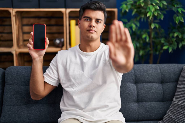 Hispanic man holding smartphone showing blank screen with open hand doing stop sign with serious and confident expression, defense gesture  - Valokuva, kuva