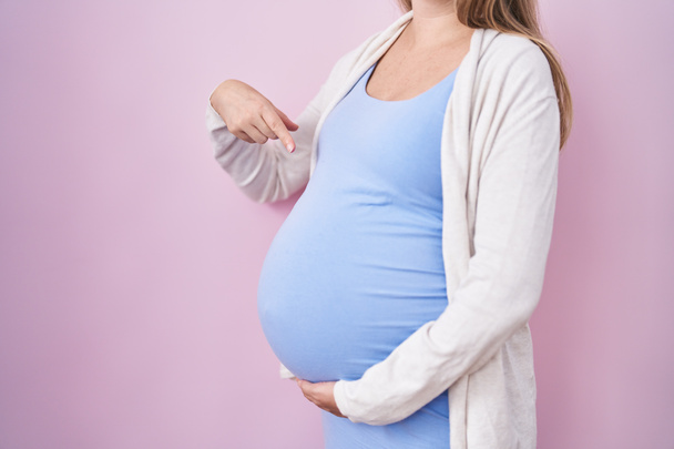 Νεαρή έγκυος γυναίκα περιμένει ένα μωρό, αγγίζοντας κοιλιά έγκυος χαμογελά χαρούμενος δείχνοντας με το χέρι και το δάχτυλο  - Φωτογραφία, εικόνα