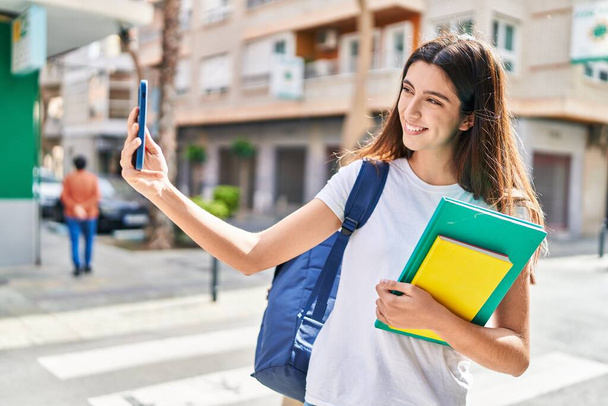 Young beautiful hispanic woman student holding books make selfie by smartphone at street - 写真・画像