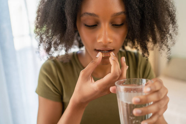 Young African girl using tablet or medical pill and holding glass of water. Healthy lifestyle, vitamin dieting. - Фото, изображение