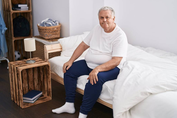 Middle age grey-haired man sitting on bed with relaxed expression at bedroom - Photo, Image