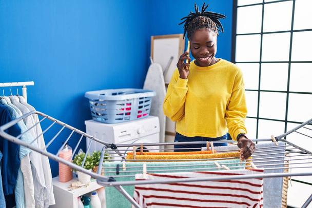 African american woman talking on the smartphone hanging clothes on clothesline at laundry room - Photo, Image