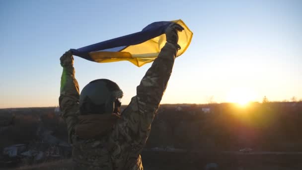 Dolly ujęcie żołnierza ukraińskiej armii podniesiony nad głową niebiesko-żółty sztandar o zachodzie słońca. Młoda dziewczyna w kamuflażu uniosła flagę Ukrainy na wsi. Koncepcja odporności na inwazję. - Materiał filmowy, wideo