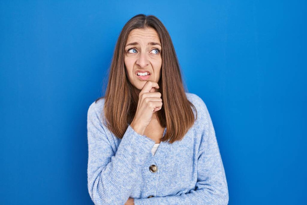Mladá žena stojí nad modrým pozadím a přemýšlí o otázce, znepokojená a nervózní s rukou na bradě  - Fotografie, Obrázek