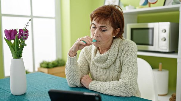 Ältere hispanische Frau mit Vapeinhalator sitzt auf Tisch und schaut sich Video auf Touchpad im Esszimmer an - Foto, Bild