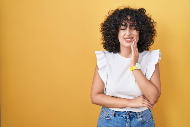 Jeune femme du Moyen-Orient debout sur fond jaune touchant la bouche avec une expression douloureuse à cause de maux de dents ou de maladies dentaires sur les dents. dentiste  - Photo, image