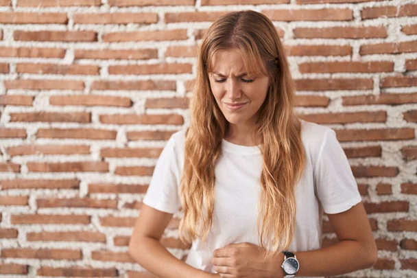 Young caucasian woman standing over bricks wall with hand on stomach because indigestion, painful illness feeling unwell. ache concept.  - Foto, Bild