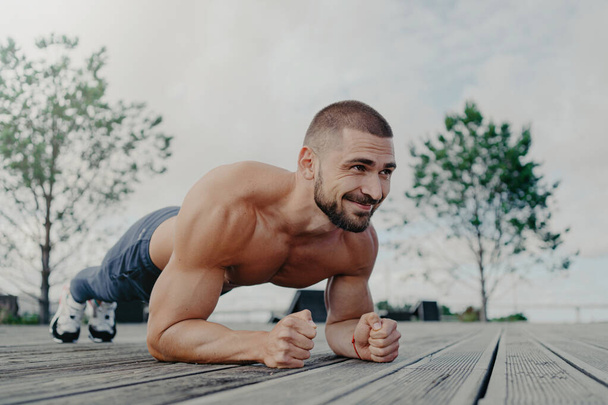Fit bearded man in plank pose, smiles outdoors. Focuses on health and fitness. - Фото, зображення