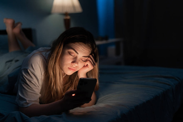 Addicted to social media young woman falling asleep with smartphone at night in bed. Mobile use addiction concept. - Photo, Image