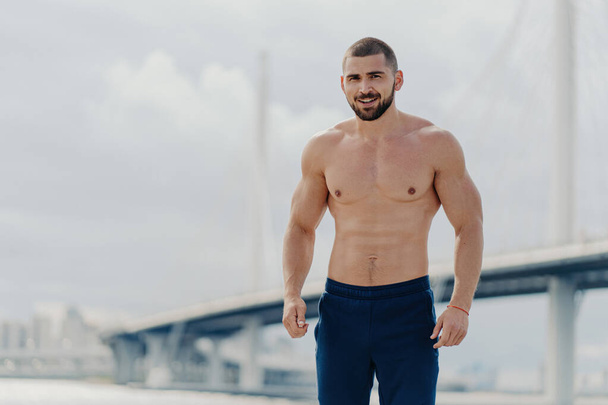 Confident bearded man with naked torso poses by river bridge, leads healthy lifestyle, has muscular body. - Photo, Image