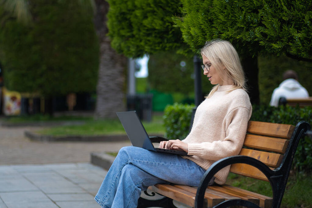 Young surprised blonde woman with glasses is looking at a laptop while sitting on a park bench. Student is sitting in the park with a laptop - Foto, immagini