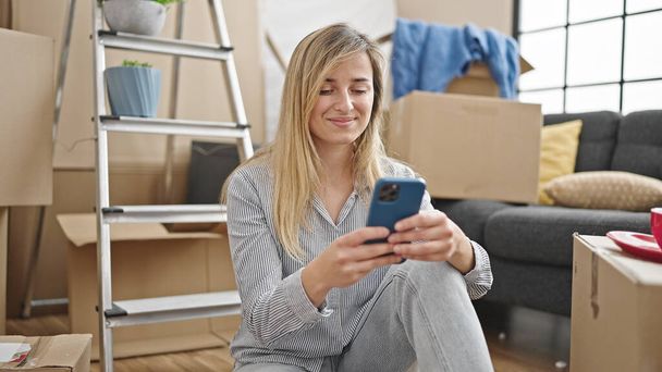 Молодая блондинка с помощью смартфона сидит на полу в новом доме - Фото, изображение