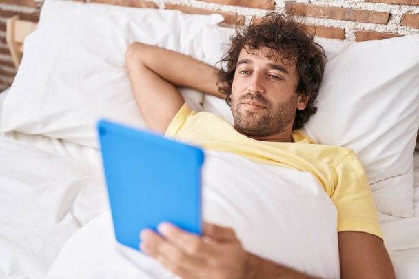 Young hispanic man watching video on touchpad lying on bed at bedroom - Foto, Imagem