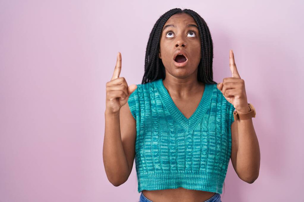 Jeune afro-américain avec des tresses debout sur fond rose étonné et surpris regardant vers le haut et pointant avec les doigts et les bras levés.  - Photo, image