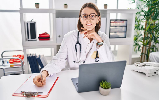 Young caucasian woman doctor using laptop writing on document at clinic - Zdjęcie, obraz