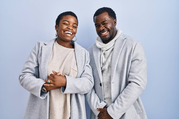Junges afrikanisch-amerikanisches Paar, das vor blauem Hintergrund steht und augenzwinkernd in die Kamera blickt, mit sexy Gesichtsausdruck, fröhlichem und glücklichem Gesicht.  - Foto, Bild