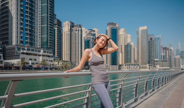 Beautiful young woman standing and Dubai marina skyline. having rest after morning run at the city. Portrait of satisfied woman after fitness exercises. - Foto, imagen