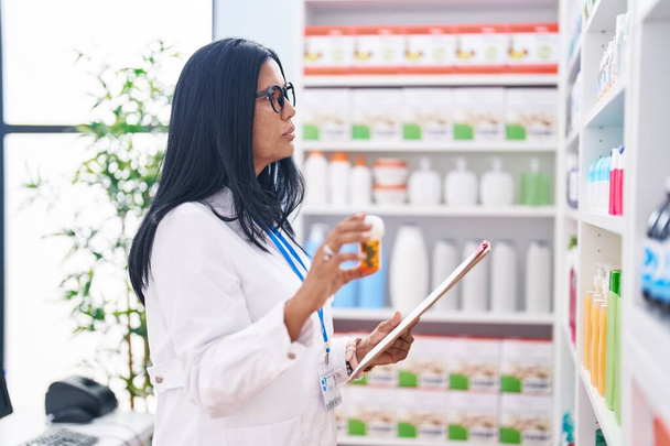 Farmacéutica hispana de mediana edad sosteniendo píldoras botella leyendo documento en farmacia - Foto, imagen