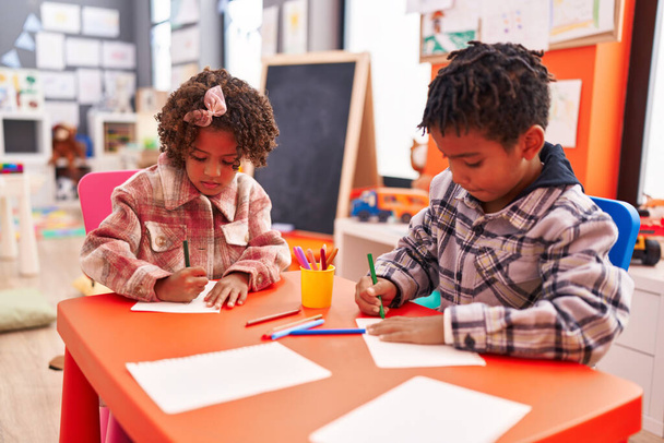 Uroczy afrykański amerykański chłopiec i dziewczynka przedszkolaków siedzi na stole rysunek na papierze w przedszkolu - Zdjęcie, obraz