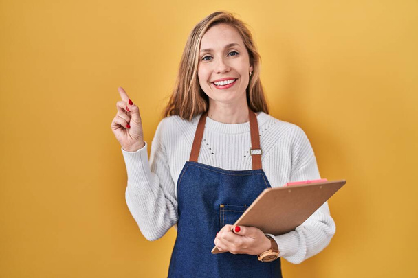 Młoda blondynka nosząca profesjonalny fartuch kelnerski trzymająca schowek uśmiechając się szczęśliwa wskazując ręką i palcem z boku  - Zdjęcie, obraz