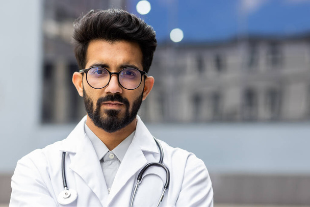Close up portret młodego indyjskiego lekarza, mężczyzna w białym płaszczu medycznym poważne i myślenie patrząc w obiektyw, pediatra poza kliniką zbliżenie. - Zdjęcie, obraz