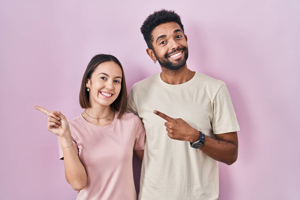 若いヒスパニック系のカップルが一緒に喜びと自然な表情で側に手と指を指して顔に笑顔で陽気にピンクの背景の上に  - 写真・画像