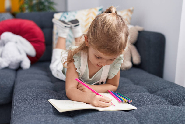 Adorable chica rubia estudiante de preescolar sentado en el sofá dibujo en el cuaderno en casa - Foto, Imagen