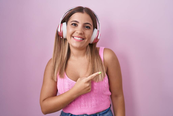Mujer rubia joven escuchando música usando auriculares alegres con una sonrisa en la cara apuntando con la mano y el dedo hacia el lado con expresión feliz y natural  - Foto, Imagen