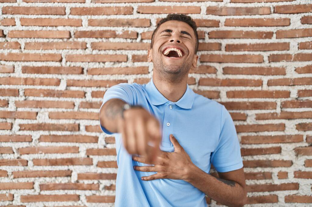 Brazilský mladík stojí nad cihlovou zdí a směje se ti, ukazuje prstem na kameru s rukou přes tělo, výraz studu  - Fotografie, Obrázek